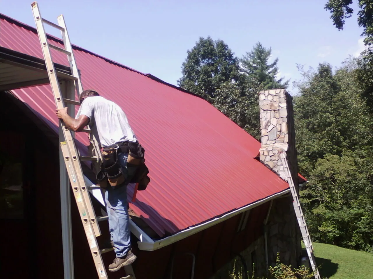 Roofer on a ladder beside a red metal roof and stone chimney — Soffit & Fascia Repair by Roofing Brothers in Grays River